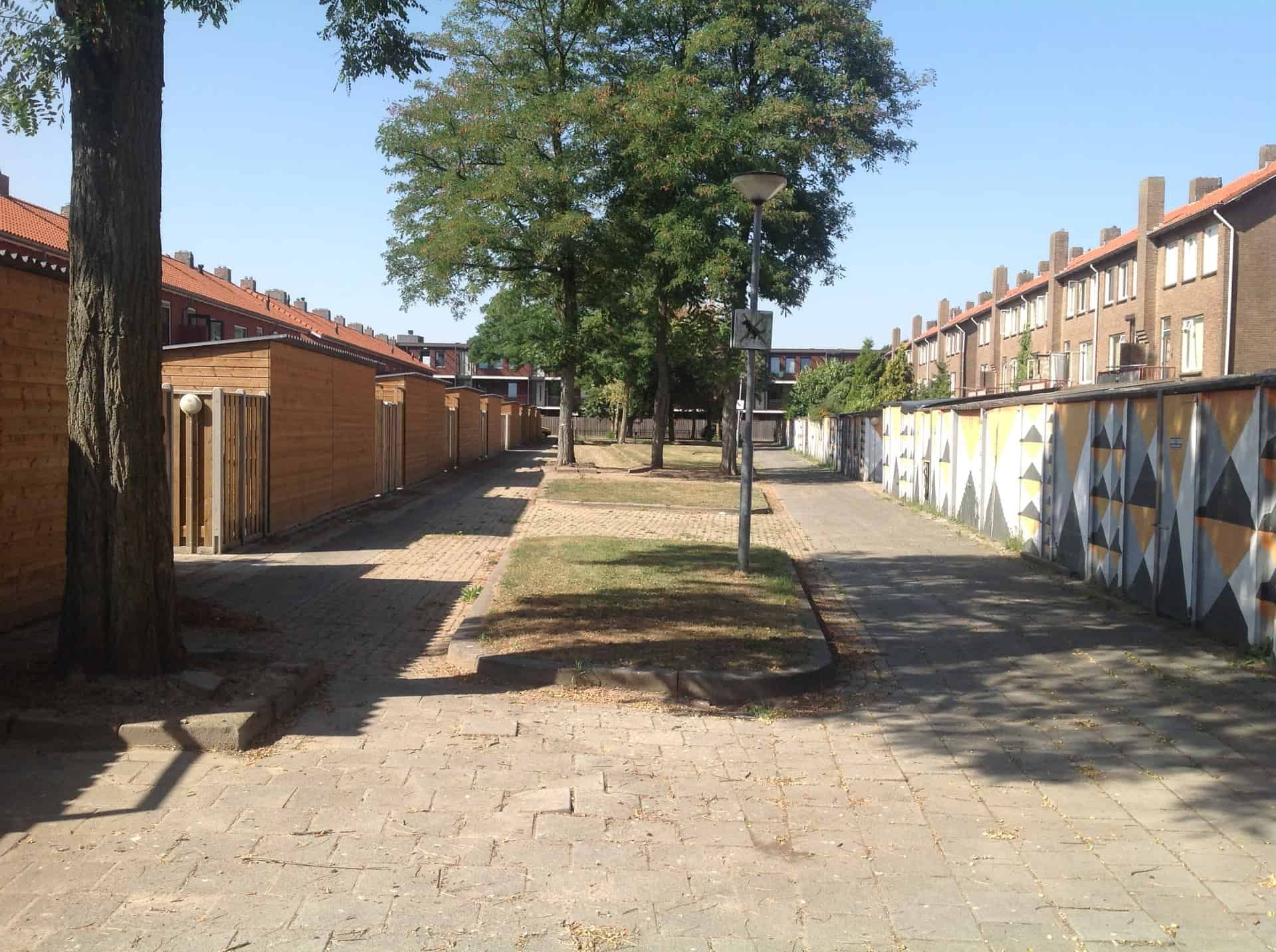 Straat met rechts oude bergingen en links nieuwe houten bergingen en tussenin bomen en een pad van stoeptegels