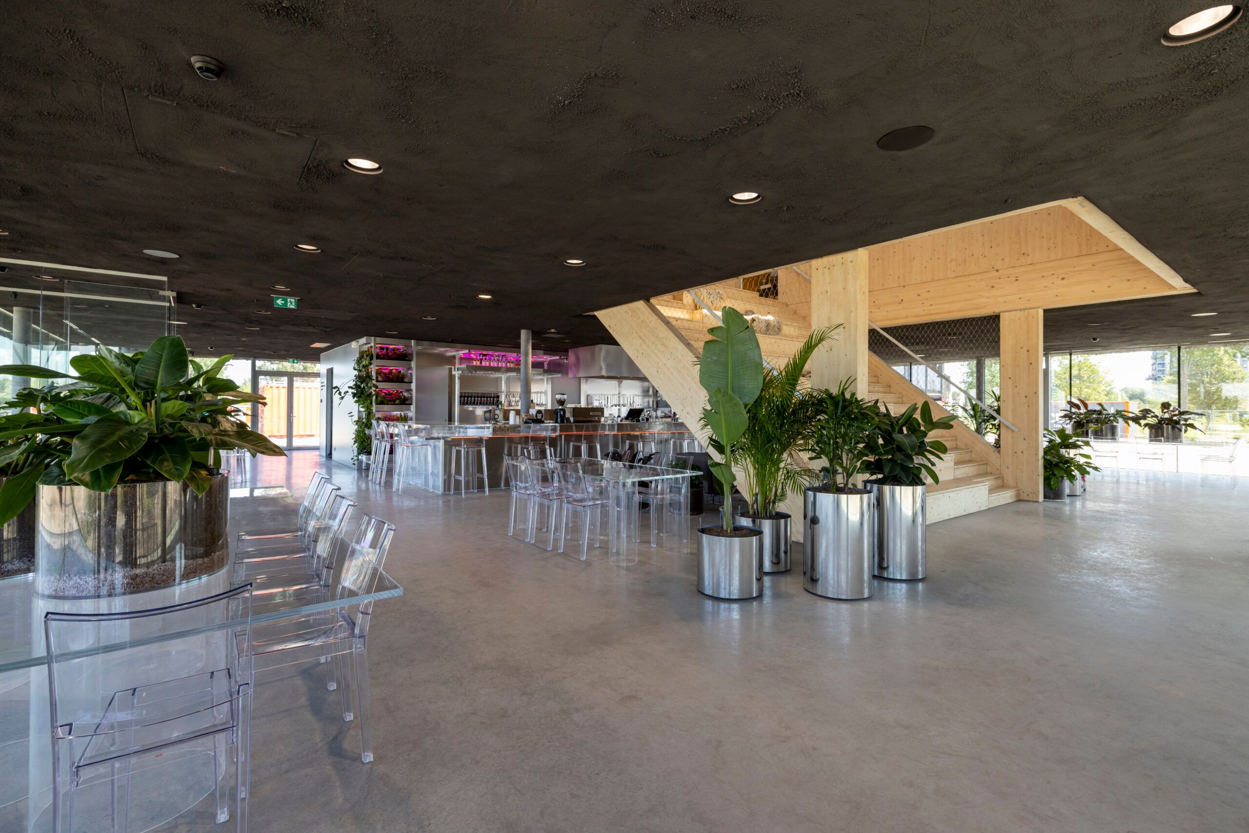 Restaurant met doorzichtig meubilair, veel planten, een bar en een houten trap