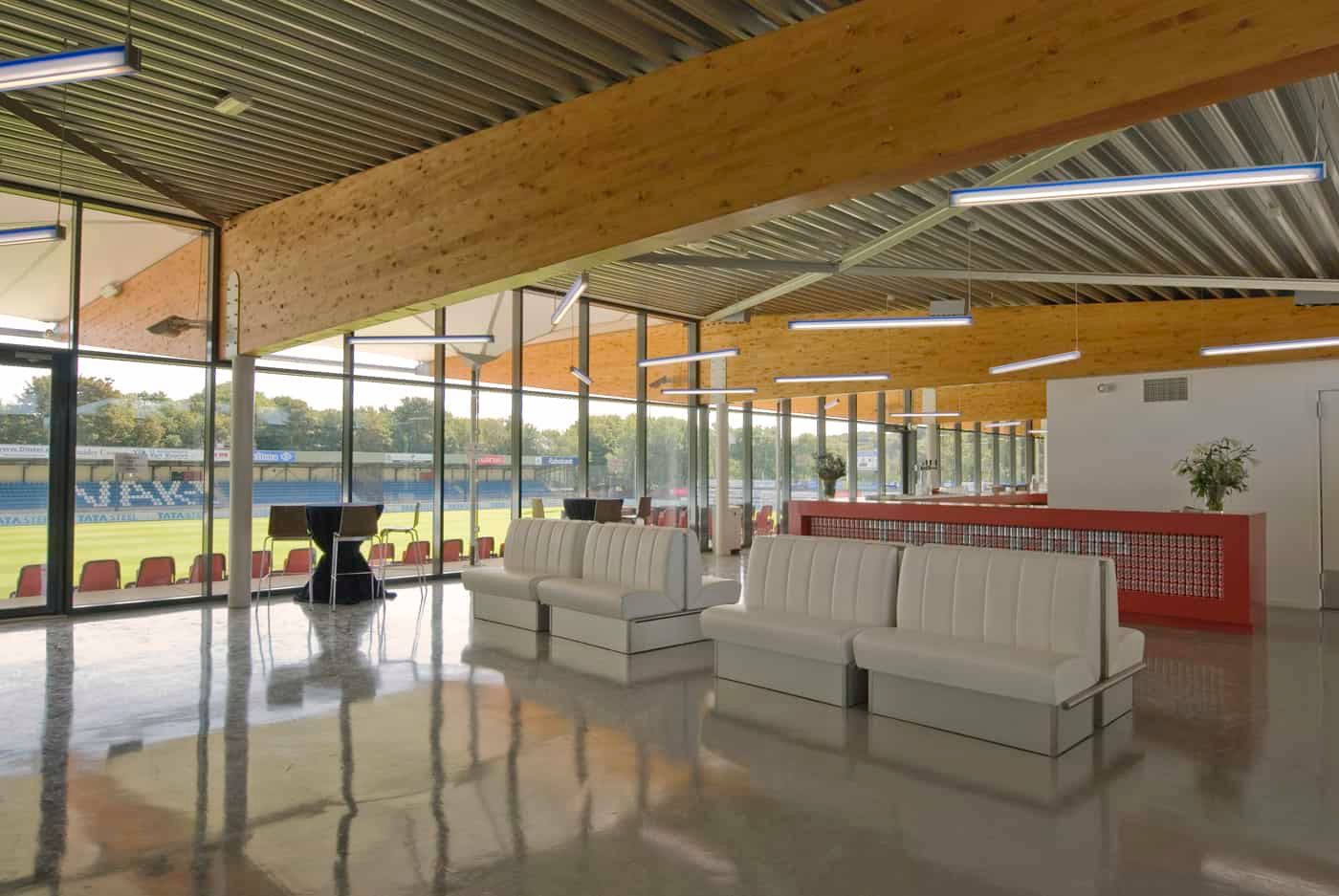 Binnenkant van tribune met houten balken aan het plafond, witte banken, een rode bar en uitzicht op het voetbalveld