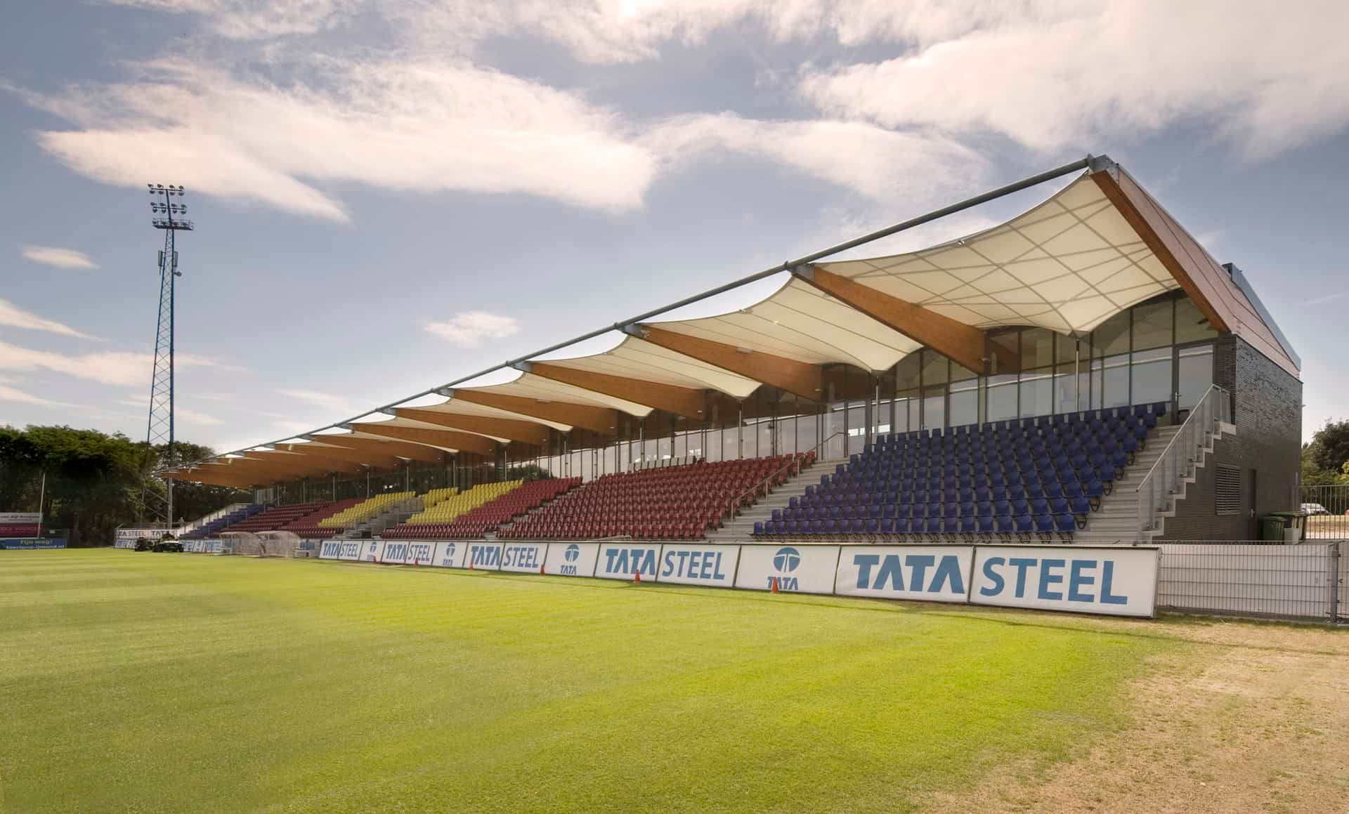 Tribune aan een voetbalveld met rode, gele en blauwe stoelen en een doek op houten balken als overkapping