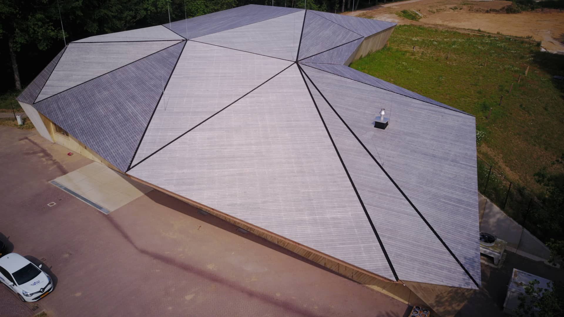 Houten gebouw met schuine grijze daken vanaf de bovenkant met een auto en grasveld