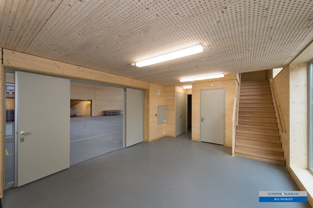 De hal van hout met houten trap, grijze deuren, ramen en uitzicht op de sportzaal