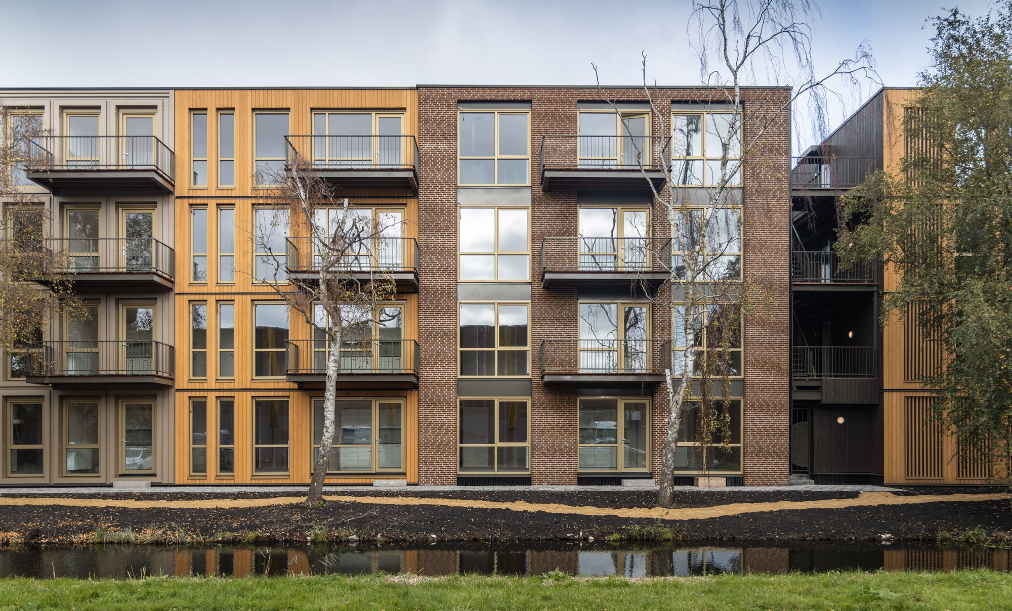 Voorkant houten met bakstenen appartementencomplex met vier woonlagen achter een beekje, bomen en een grindpad ervoor
