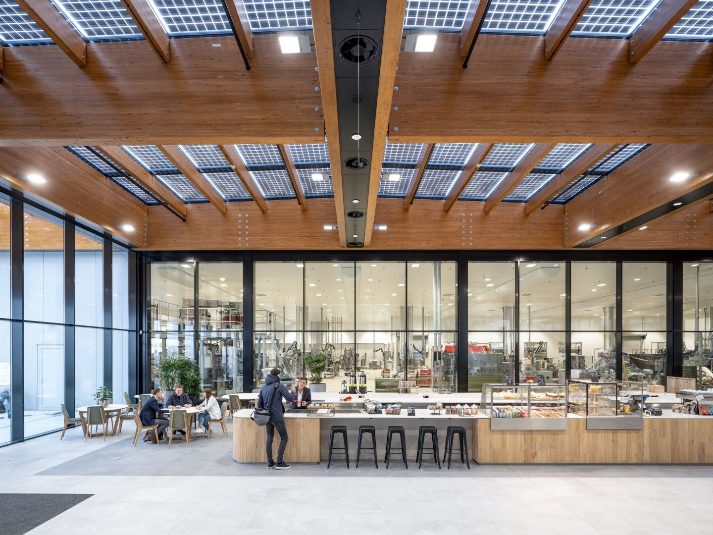 De houten bar in de kantine met barkrukken, stoelen en tafels, een houten plafond met zonnepanelen en grote ramen met zwarte kozijnen