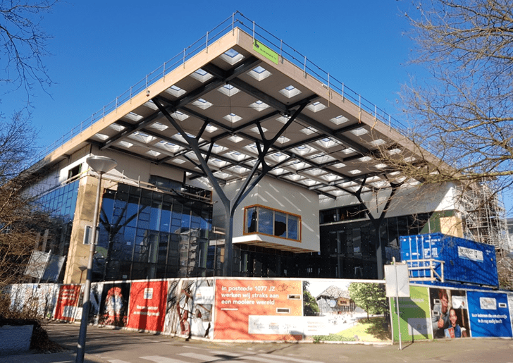 Glazen gebouw van voren met overkapping in aanbouw van hout met vierkante gaten in het dak
