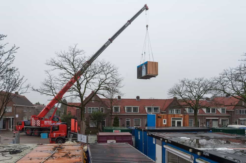 Een lichtbruine box die wordt vervoerd via een rode, hoge kraan