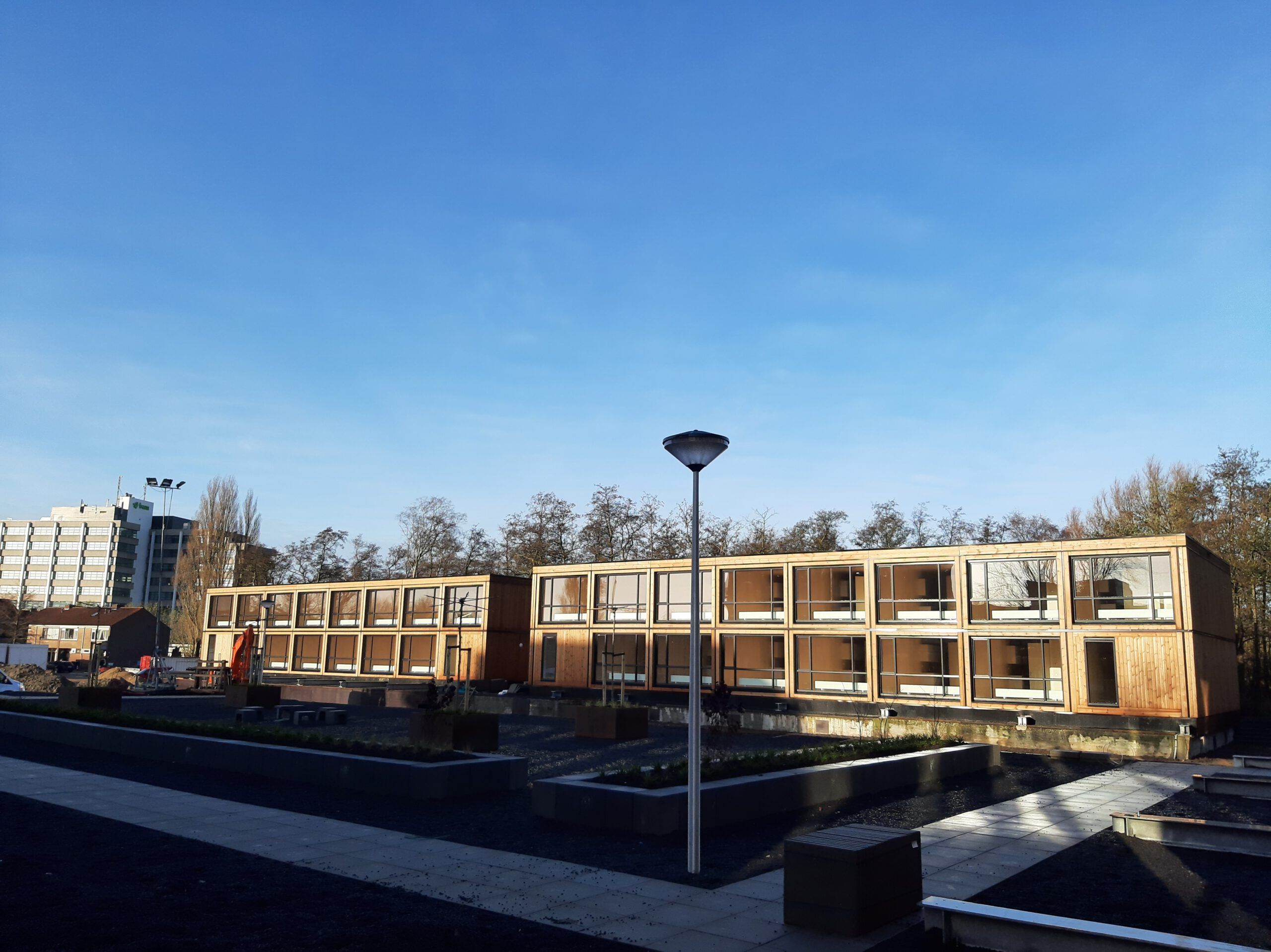 Houten gebouw met grote ramen voor bomen en grind, een pad van stoeptegels en een lantaarnpaal ervoor