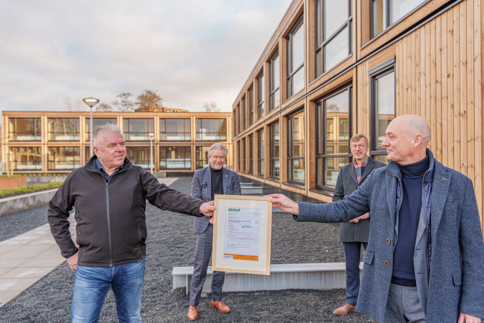 Vier mannen met een certificaat in handen voor een houten woongebouw