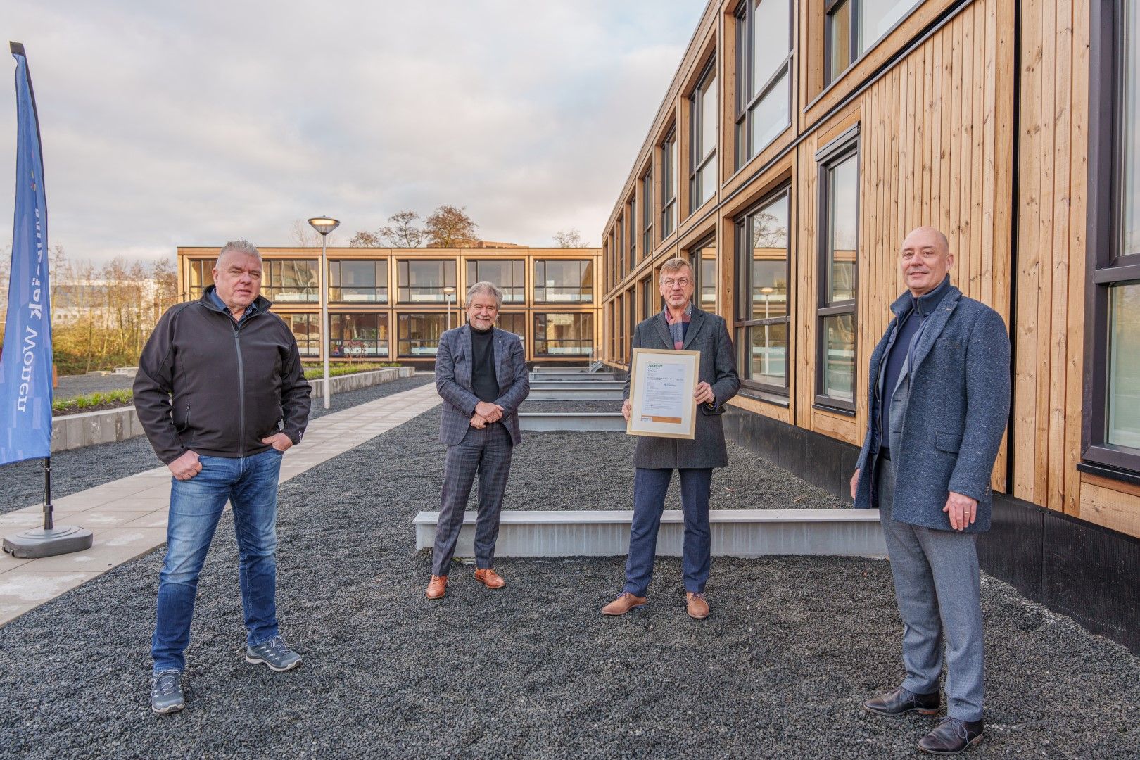 Vier mannen waarvan één een certificaat vast heeft op een grindbak voor houten woongebouwen