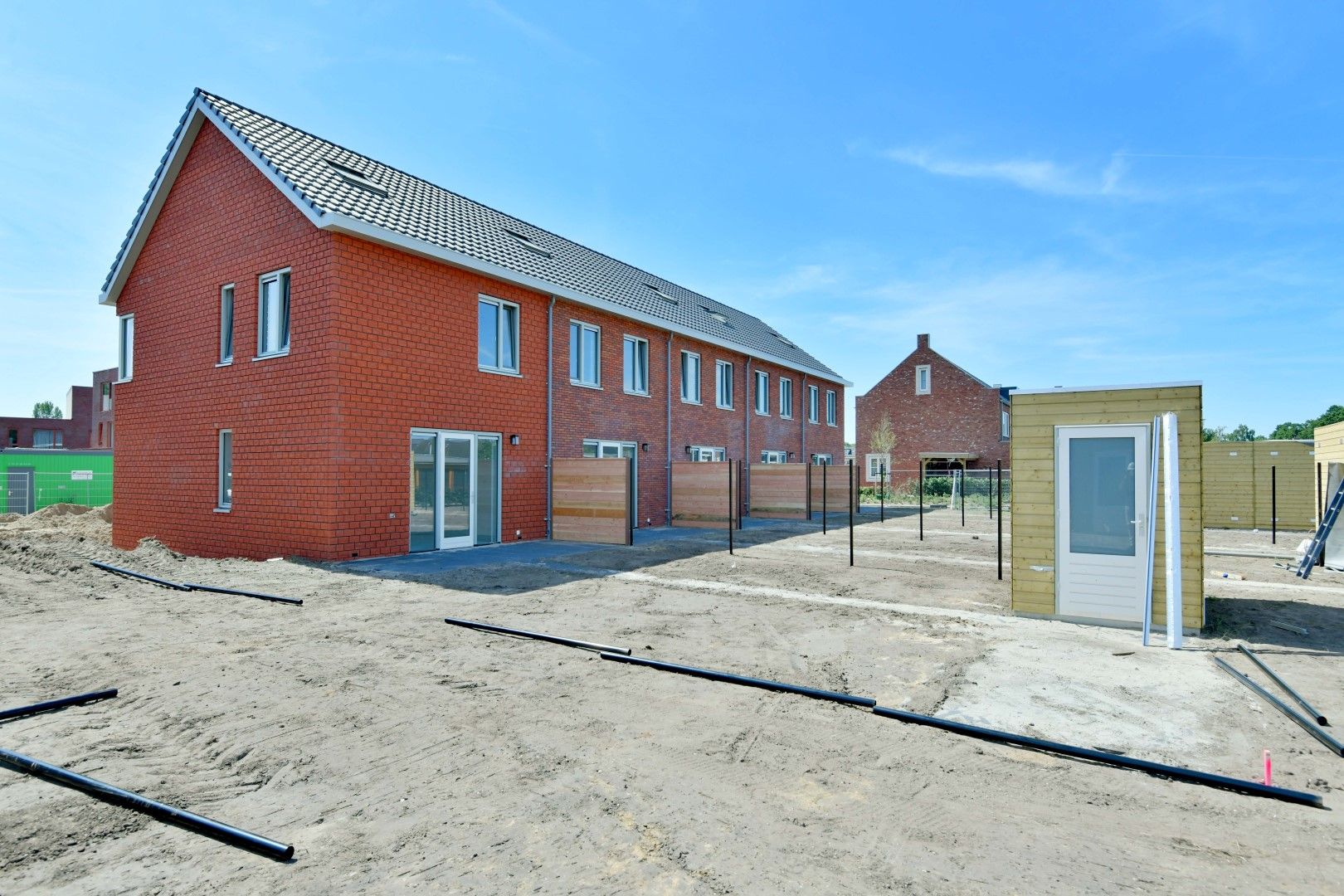 Rode nieuwbouw rijtjeshuizen met zwarte daken met gele, houten berging in tuin.
