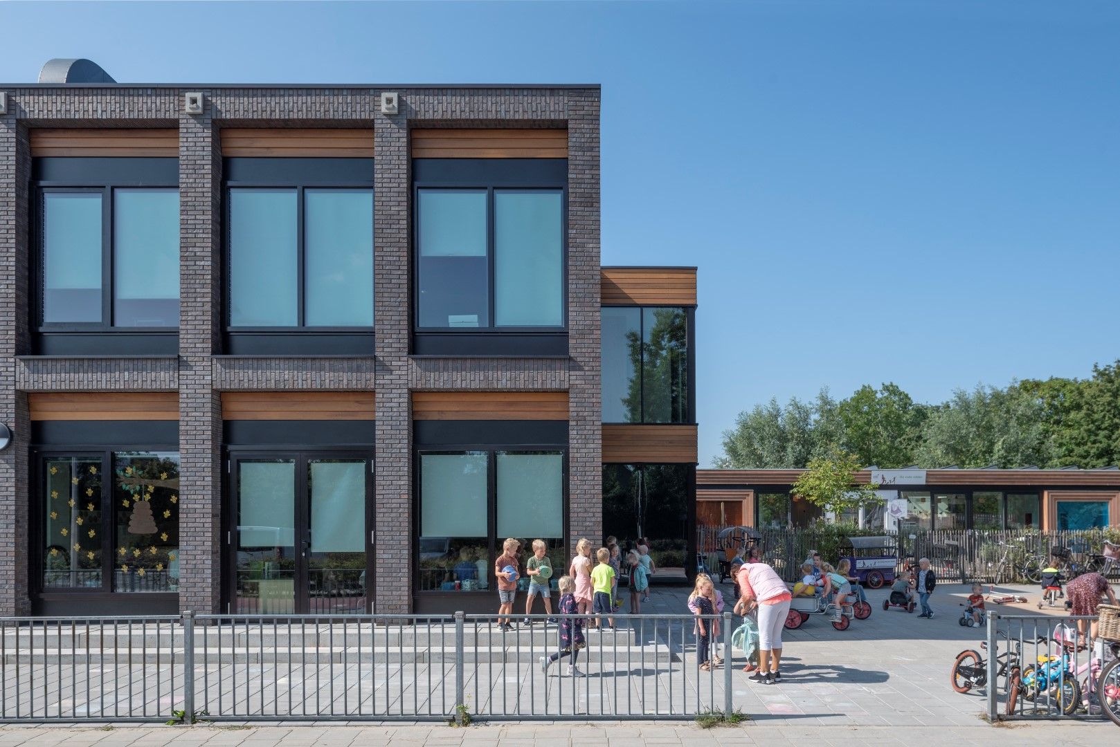 Grijs gebouw met houten elementen, grote ramen en een plein met spelende kinderen