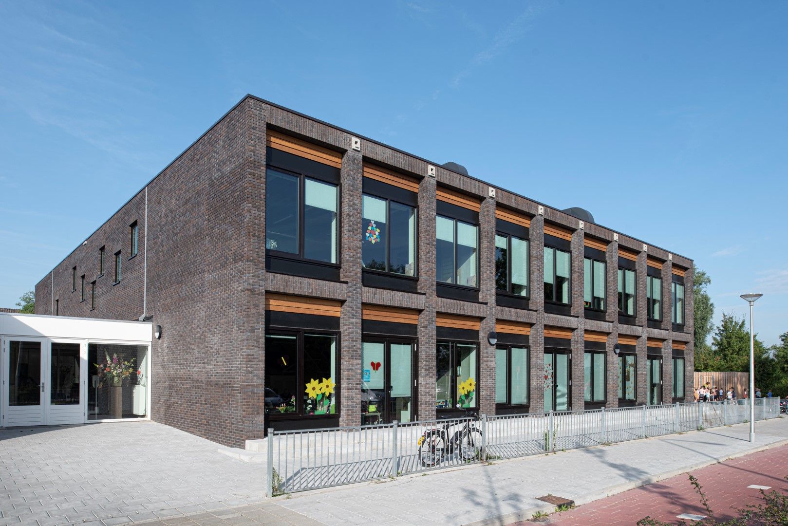 Een vierkant grijs gebouw met grote ramen en houten balken boven de ramen vanaf de straatkant met fietspad en een plein