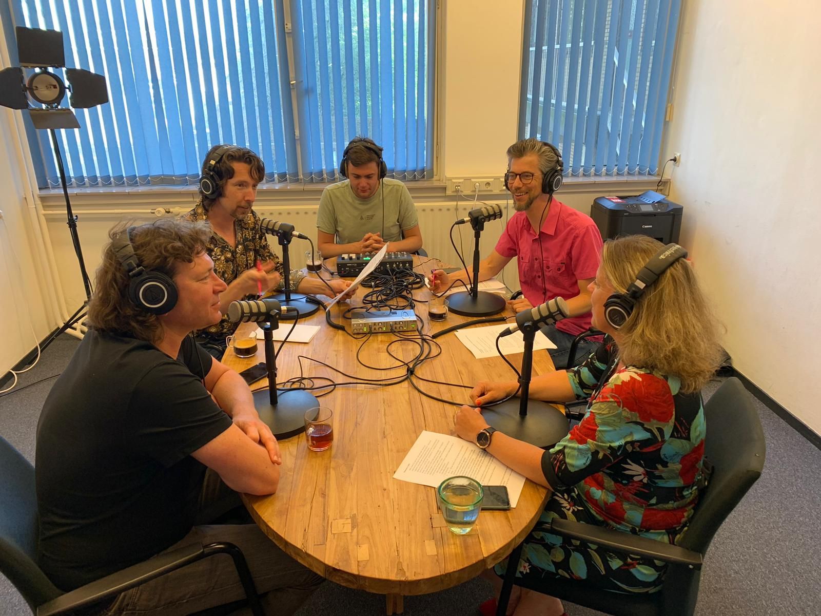 Vijf mensen zittend aan een houten tafel met microfoons tijdens een podcastopname