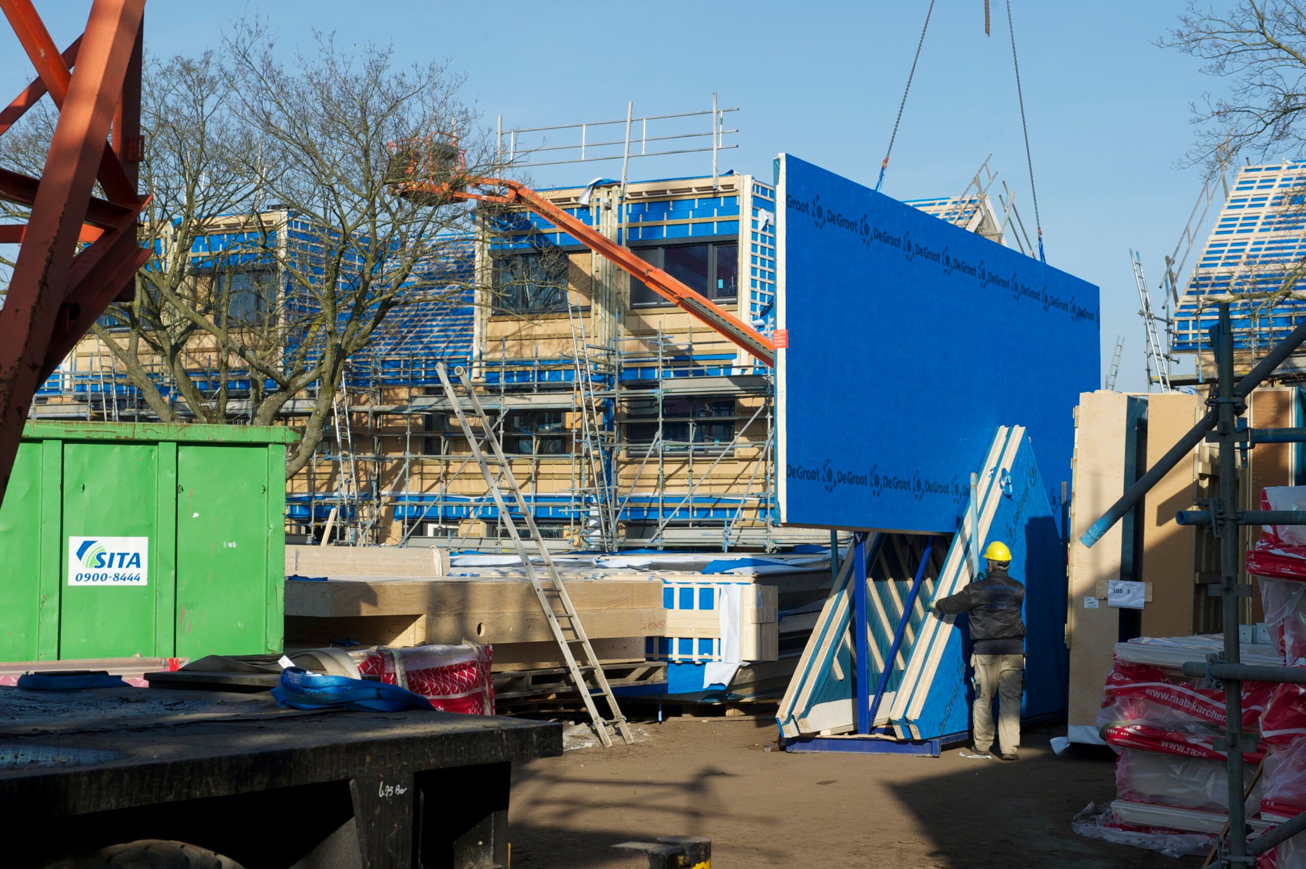 Gebouw in aanbouw met hout en blauwe elementen en een groene container ervoor