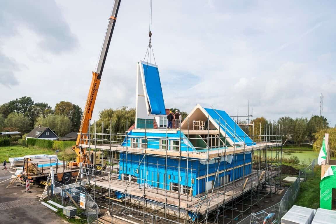Een huis in aanbouw met steigers om de woning heen en een hijskraan die een wand optilt
