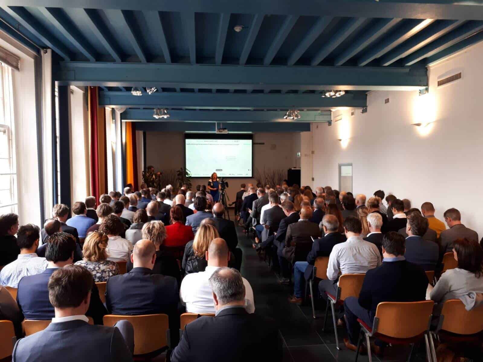 Grote groep mensen die zitten in een zaal met blauw plafond