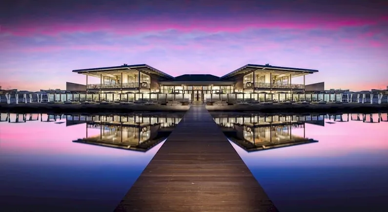 Gebouw met veel ramen en een houten steiger aan het water met roze lucht