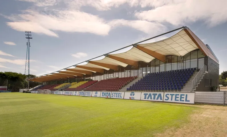 Tribune aan een voetbalveld met rode, gele en blauwe stoelen en een doek op houten balken als overkapping