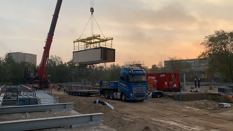 Bouwplaats van zand met houten module aan een hijskraan met een vrachtwagen eronder