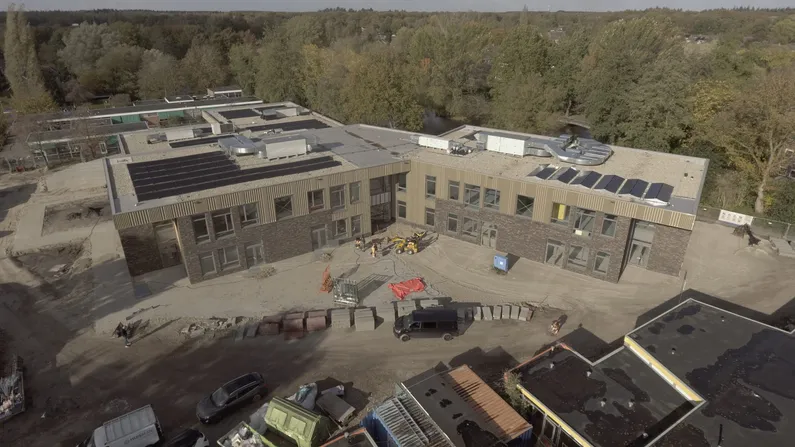 Houten school in aanbouw op een zandoppervlakte met veel bomen