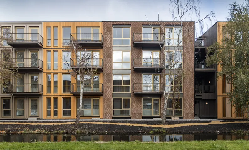 Voorkant houten met bakstenen appartementencomplex met vier woonlagen achter een beekje, bomen en een grindpad ervoor
