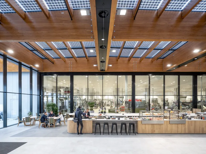 De houten bar in de kantine met barkrukken, stoelen en tafels, een houten plafond met zonnepanelen en grote ramen met zwarte kozijnen