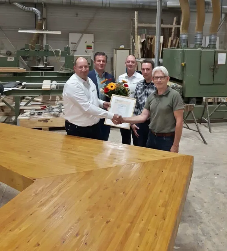 Vijf mensen aan een houten tafel met een certificaat en een bloemetje in hun handen
