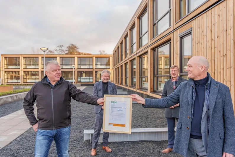 Vier mannen met een certificaat in handen voor een houten woongebouw