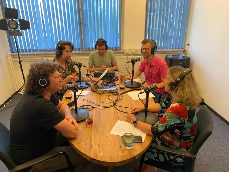 Vijf mensen zittend aan een houten tafel met microfoons tijdens een podcastopname