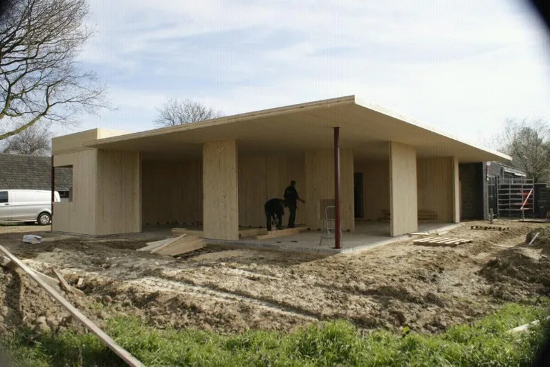 Houten gebouw op zand en gras waar twee mensen aan het werk zijn